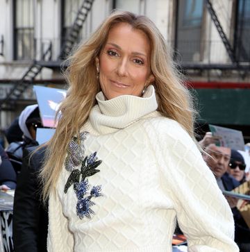 celine dion stands outside and smiles at the camera, she is wearing a white sweater with a larger ribbed collar and blue floral embrodiery