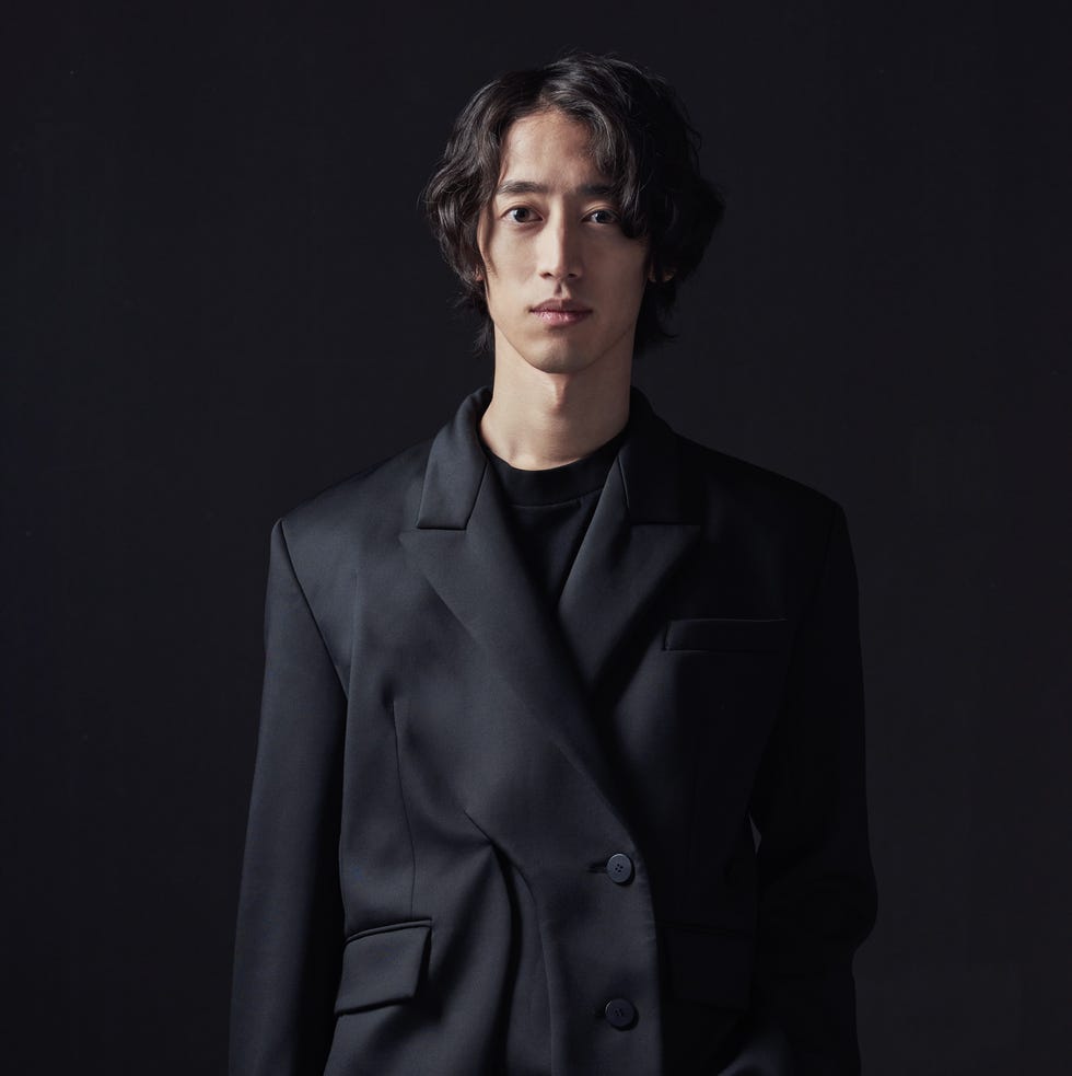 a person wearing a black oversized suit jacket stands against a dark backdrop