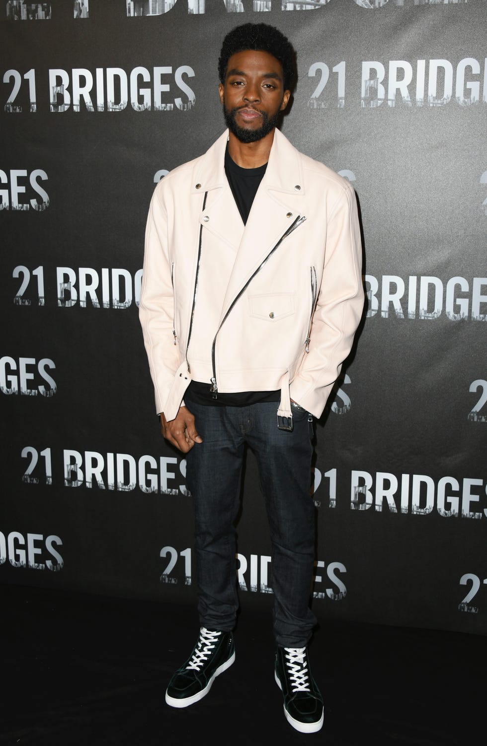 chadwick boseman stands in front of a black 21 bridges movie poster, he wears a cream leather jacket, dark jeans, and black sneakers