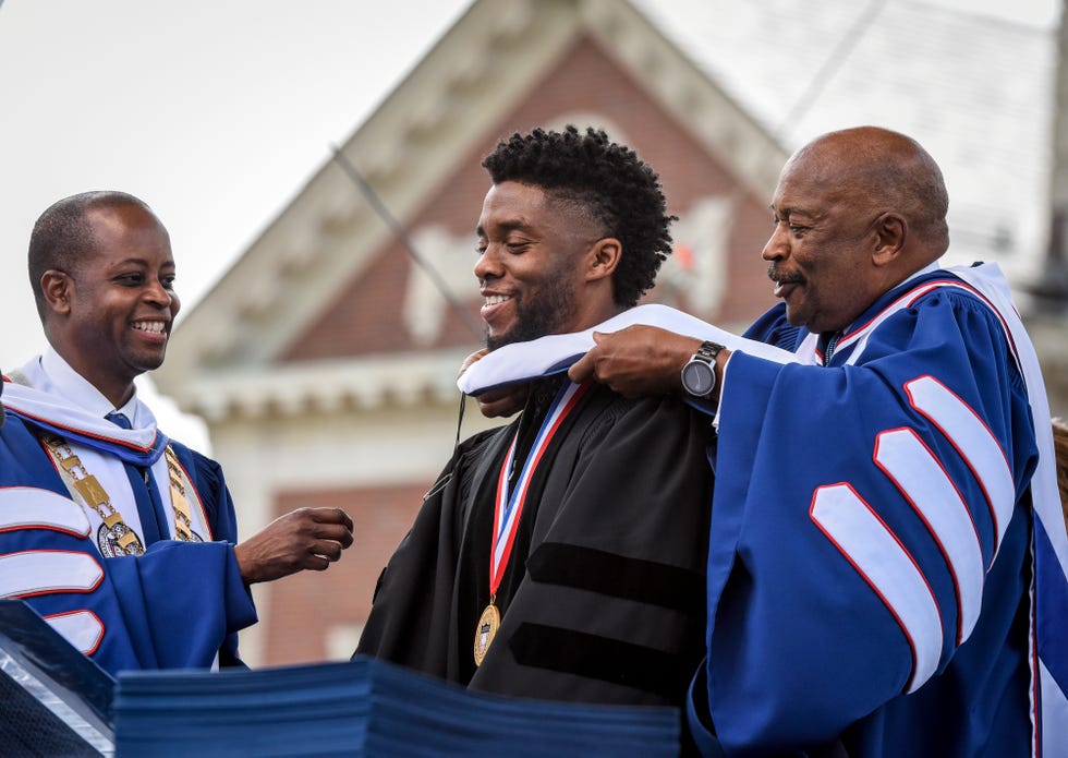 chadwick boseman stands in front of a podium in a black graduation gown, he smiles as a man places a sash on his shoulders