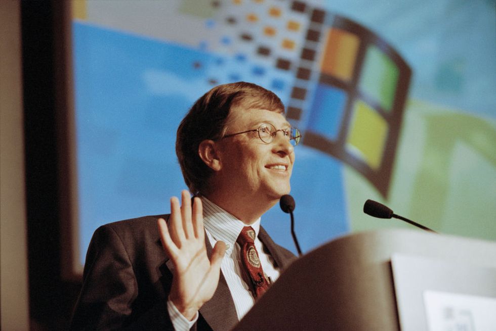 bill gates speaks at a podium with microphones, a windows logo is projected on a screen behind him