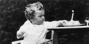 charles lindbergh jr sits on a chair and reaches to a birthday cake with a lit candle to the right