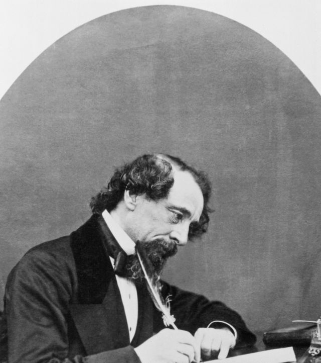 charles dickens sitting at a desk, he holds a quill above a piece of paper and looks down, he wears a suit