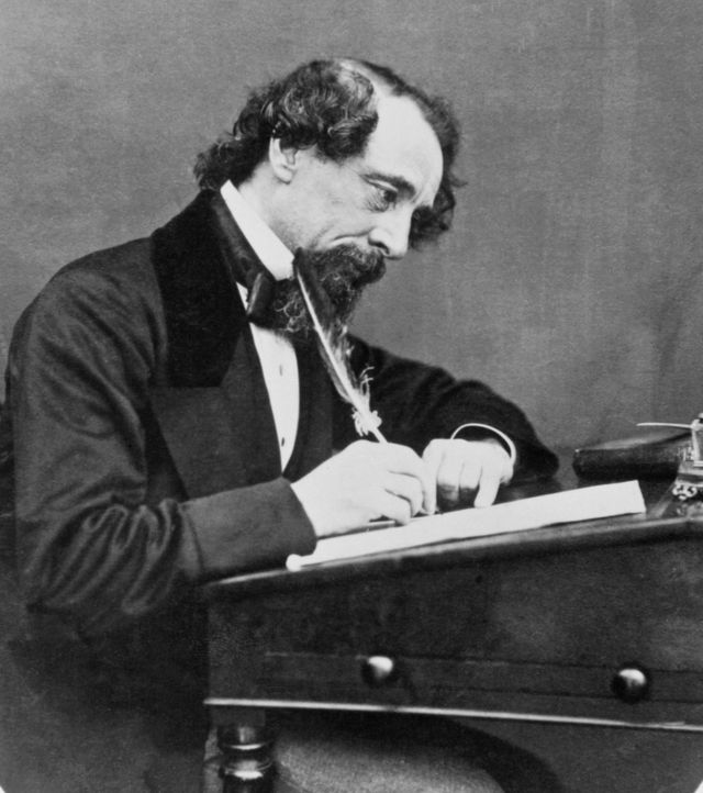 Charles Dickens sta la un birou, tine o pana deasupra unei foi de hartie si se uita in jos, poarta un costum