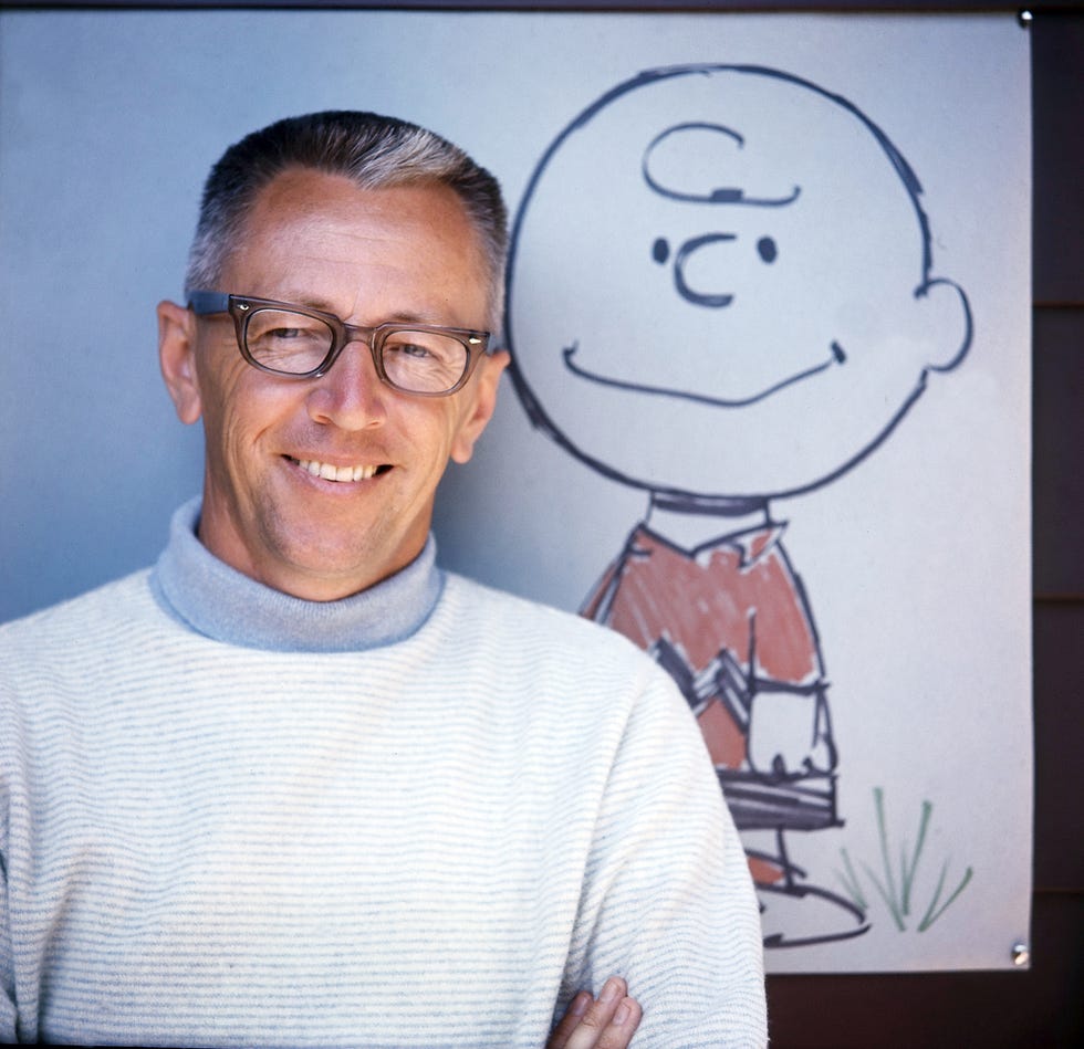 charles schulz, wearing a white and gray turtleneck and glasses, smiles while standing in front of a drawing of charlie brown