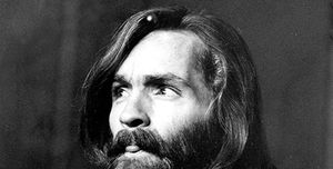 charles manson looks to the left of the camera and slightly sticks out his tongue, he wears a denim shirt and black necklace, he has shoulder length hair and a bushy full beard