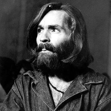 charles manson looks to the left of the camera and slightly sticks out his tongue, he wears a denim shirt and black necklace, he has shoulder length hair and a bushy full beard