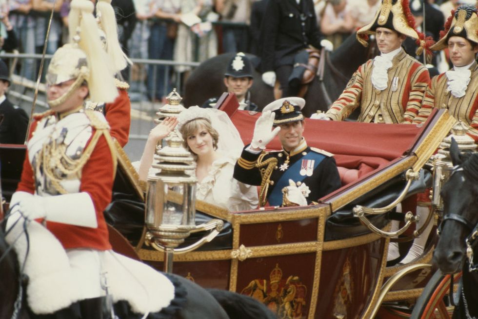 princess diana and prince charles sit in an open carriage and wave and smile at crowds, she wears a veil and wedding dress, he wears a military uniform including a hat and white gloves