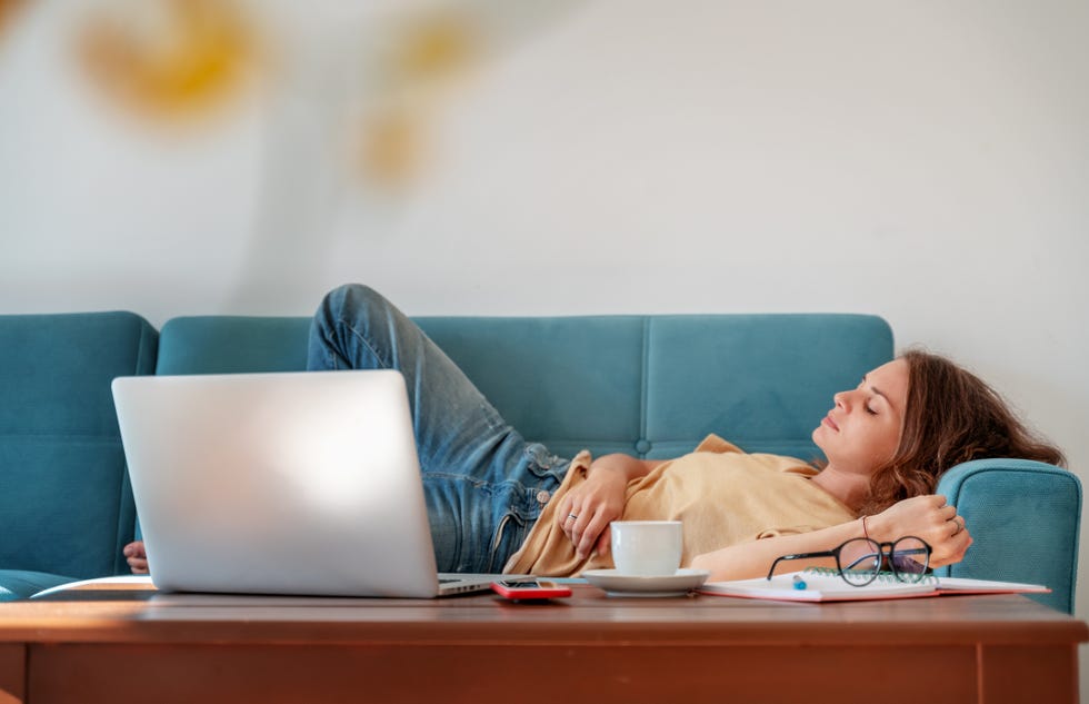 charming beautiful young girl in glasses with curly hair fell asleep on a blue sofa at home in front of a laptop, notepad and a cup of coffee, remote work and education fatigue, deadline