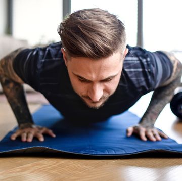 tattooed man doing push ups on yoga mat in living room with water bottle and dumbbells