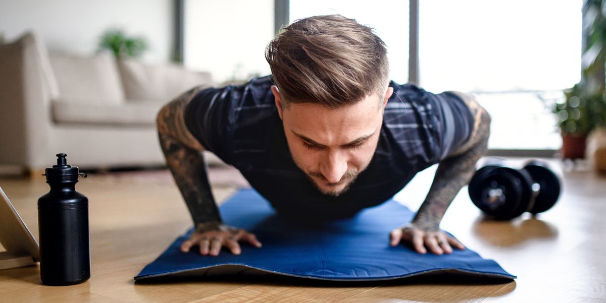 tattooed man doing push ups on yoga mat in living room with water bottle and dumbbells