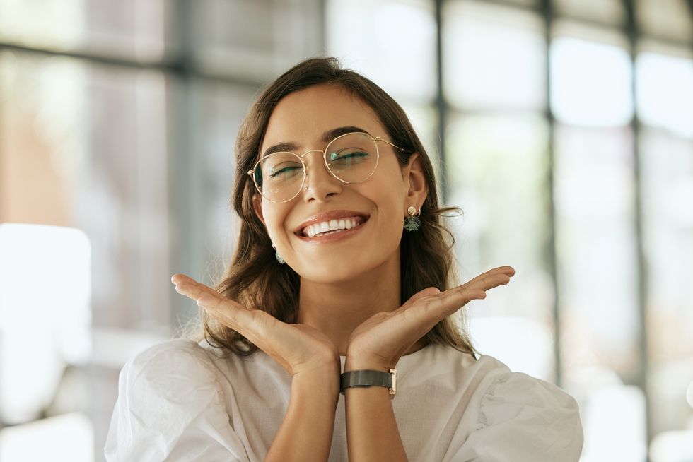 cheerful business woman with glasses posing with her hands under her face showing her smile in an office playful hispanic female entrepreneur looking happy and excited at workplace