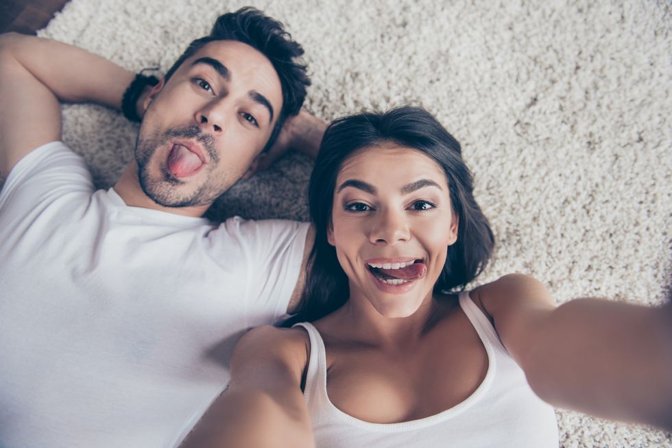 Cheerful playful hispanic sister and brother are taking selfie and making funny grimaces. They lie on the floor on beige carpet in white casual outfits indoors at home