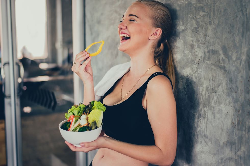 cheerful woman eating salad while standing by wall