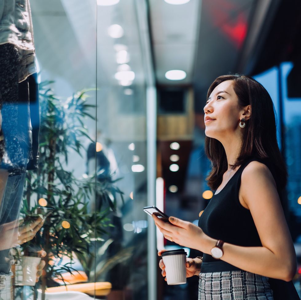 cheerful woman holding a cup of coffee, checking her smartphone while standing outside a boutique looking at shop window in the evening in the city