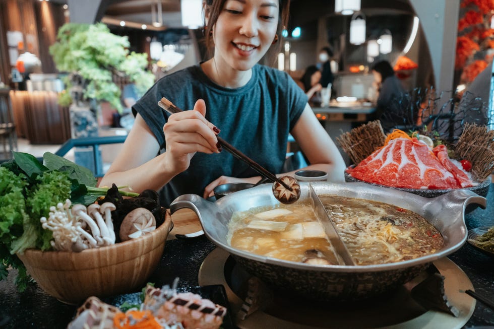 cheerful young asian woman enjoying traditional chinese hotpot with assorted fresh and scrumptious ingredients in restaurant she is boiling shiitake mushroom into the soup chinese culture chinese cuisine and food eating out lifestyle