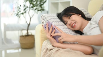 cheerful young asian woman lying on sofa and using mobile phone