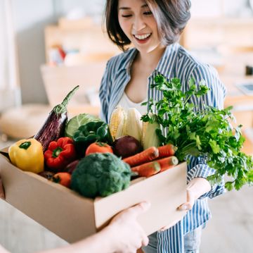 cheerful young asian woman receiving a box full of colourful and fresh organic groceries ordered online from delivery person at home