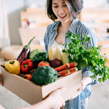 cheerful young asian woman receiving a box full of colourful and fresh organic groceries ordered online from delivery person at home