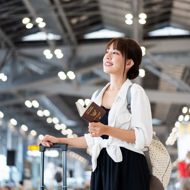 cheerful young asian woman standing in front of the flight schedule in the airport departure hall