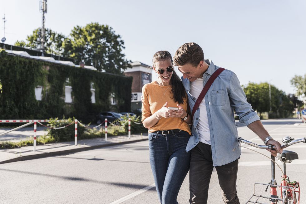 cheerful young couple looking at smartphone while walking on street in city