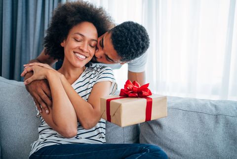 shot of a loving husband giving his wife a gift boyfriend surprise his beautiful girlfriend with present while she is sitting on the sofa in the living room at home