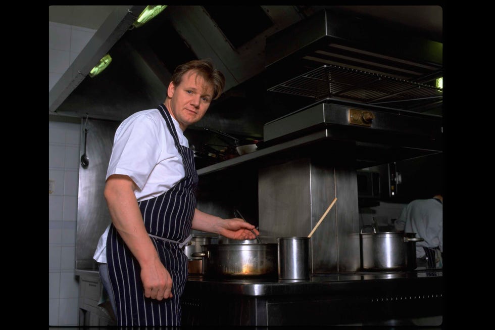 gordon ramsay stands at a stove in a kitchen, he wears a white shirt and a black and white striped apron