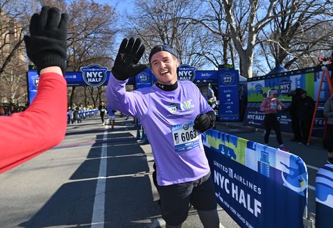 daniel humm prepares to give someone a high five at the new york city half marathon, he is wearing a purple shirt, backwards cap, black gloves, and dark bottoms
