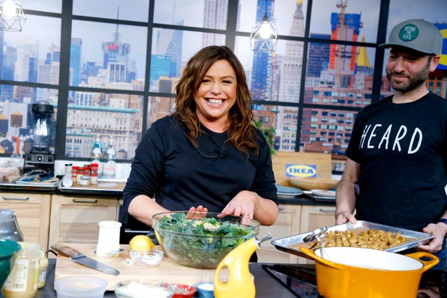 rachael ray smiles to the camera during a cooking demonstration event, she is standing in a kitchen with her hands touching a glass bowl full of salad greens she is wearing a a navy long sleeve shirt and her microphone wire is just visible, behind her are glass windows and a cityscape in the background