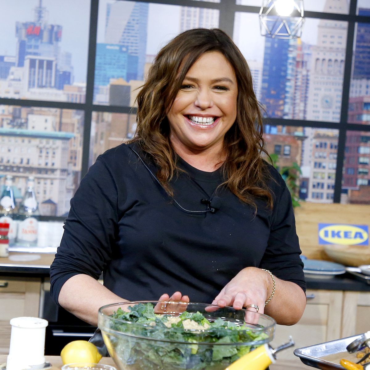 rachael ray smiles to the camera during a cooking demonstration event, she is standing in a kitchen with her hands touching a glass bowl full of salad greens she is wearing a a navy long sleeve shirt and her microphone wire is just visible, behind her are glass windows and a cityscape in the background