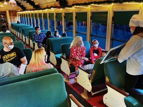 chef on an old fashioned train with green velvet seats holding up a story book in front of a little girl and a woman in pajamas there is a family in the aisle next to them and in back of them a few rows back everyone is masked