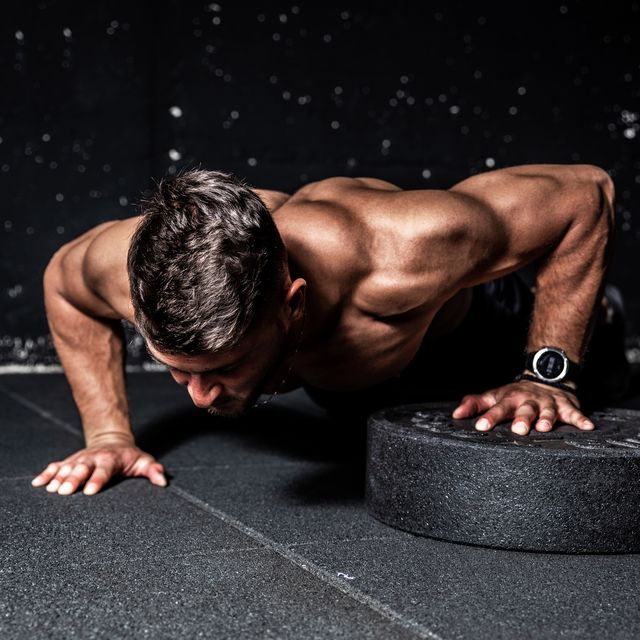 push ups, young strong sweaty focused fit muscular man with big muscles performing push ups with one hand on the barbell weight plate for training hard core workout in the gym