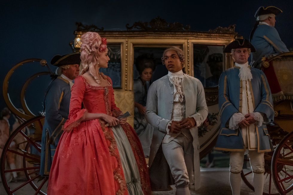 uma foto promocional do filme chevalier, apresentando os atores lucy boynton e ﻿kelvin harrison jr em trajes do século 18, em frente a uma carruagem dourada atendida por várias pessoas em uniformes azuis