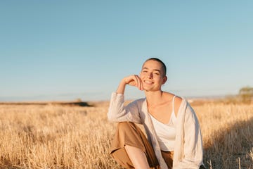 close up image of young woman looking at camera, with short hair, sitting with legs half crossed and arm resting on her leg, in cheerful attitude, laughing dressed in casual clothes in light tones in the middle of a meadow at sunset in the countryside moment of enjoyment and relaxation on a summer evening
