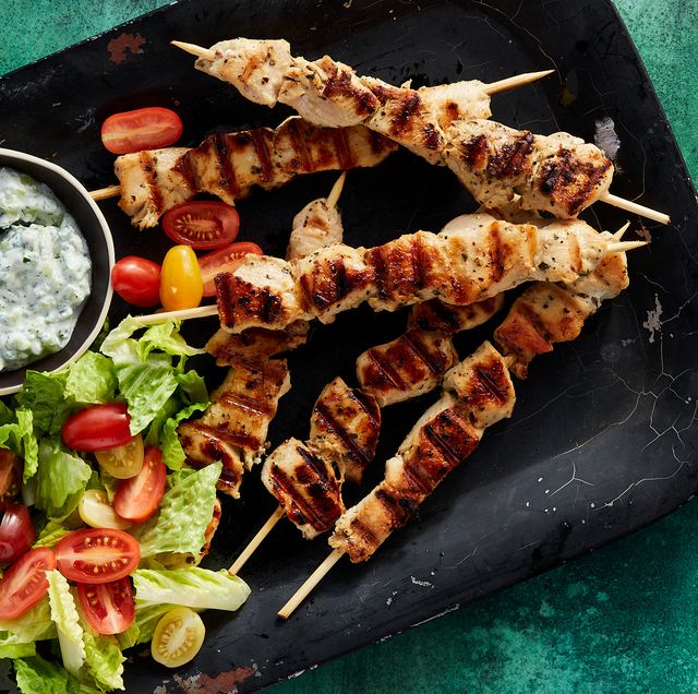 chicken souvlaki skewers surrounded by a bowl of tzatziki, cherry tomatoes, and chopped romaine on a black platter on a dark green background