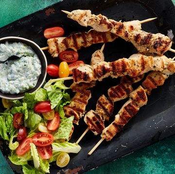 chicken souvlaki skewers surrounded by a bowl of tzatziki, cherry tomatoes, and chopped romaine on a black platter on a dark green background