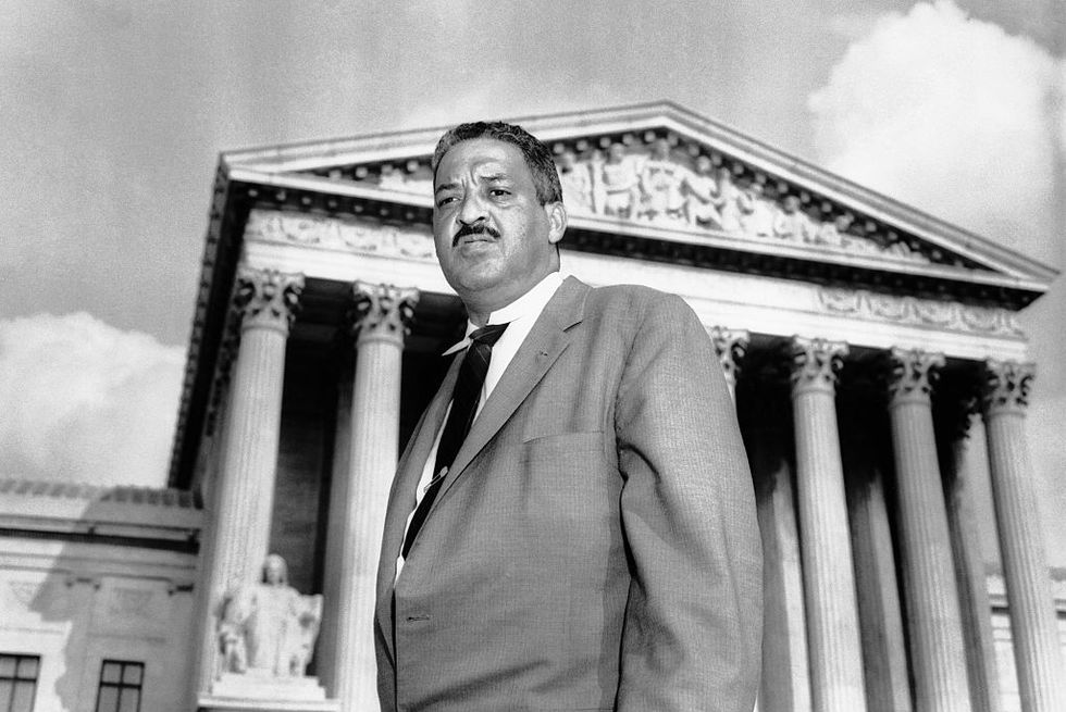 thurgood marshall stands in a suit and tie outside the us supreme court building, the building has several tall columns and ornate sculptures