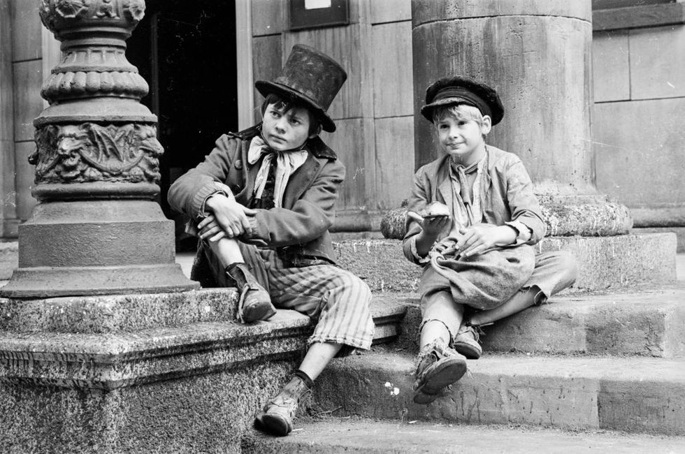 a black and white publicity still from the film oliver featuring two young actors in period costumes sitting on stone steps and looking off camera