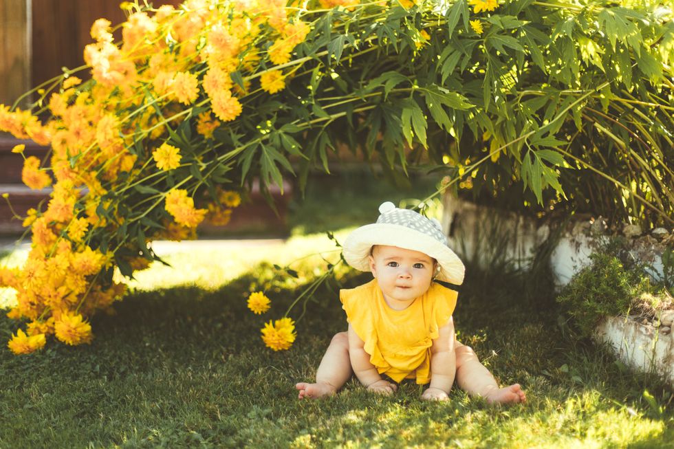a baby girl in a sun hat is sitting in the garden under a large yellow bush of flowers