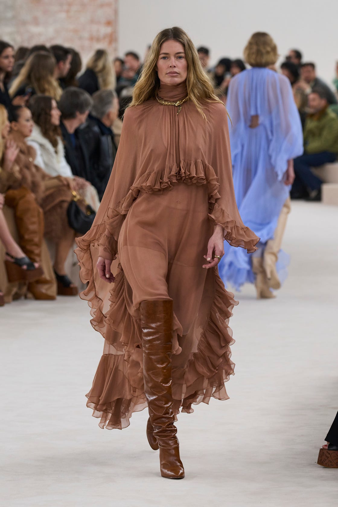 Model walking on runway wearing a flowing ruffled brown dress with kneehigh brown boots