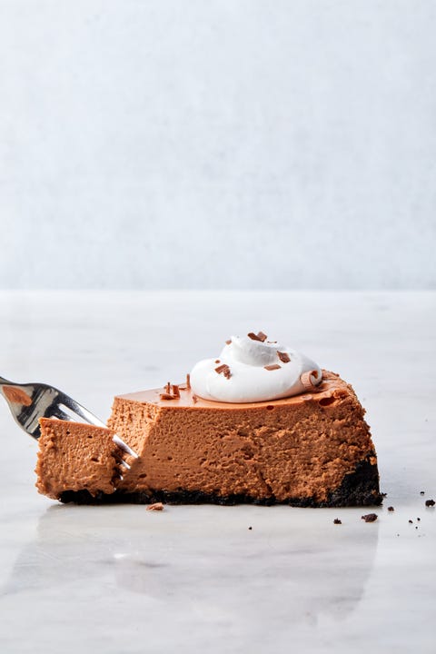 Chocolate Cheesecake slice of chocolate cheesecake with oreo crust, topped with whipped cream and chocolate curls, with a fork taking a bite out