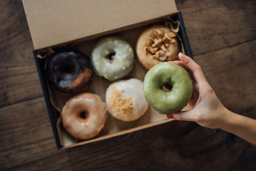 una mano sostiene un donut sacado de una caja donde hay más con diferentes glaseados