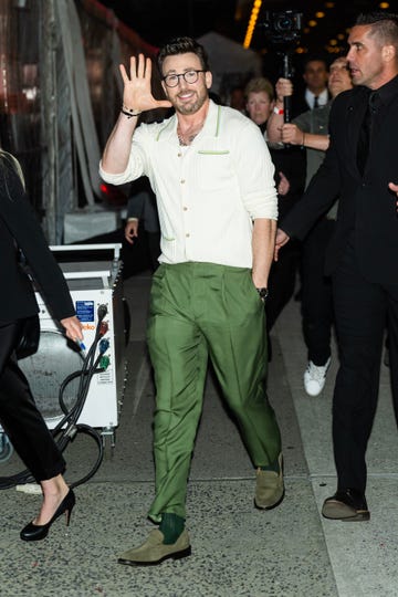 chris evans waves and smiles while walking on a city sidewalk, he wears a white button up sweater with green trim, green pants, and circular glasses