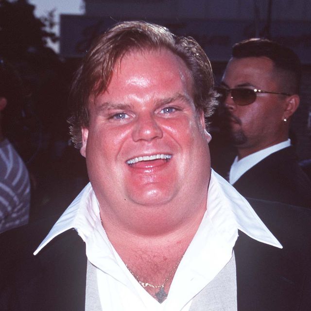 chris farley smiles at the camera, he is wearing a dark suit jacket, a white collared shirt that is unbuttoned at the neck and a silver necklace