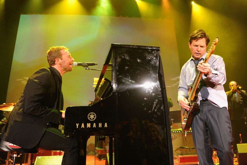 chris martin plays a black upright piano and sings into a microphone while michael j fox stands to the right and plays electric guitar