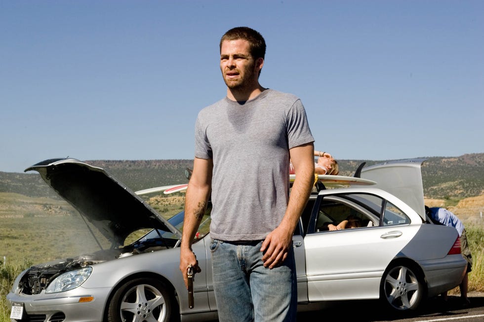 chris pine holding a gun next to a broken down car on a rural road in carriers