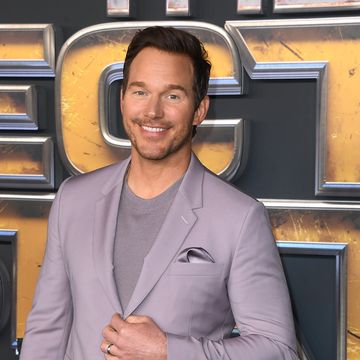 a man smiles at the camera while standing and holding his lavender suit jacket together with one hand, he also wears a gray sweater