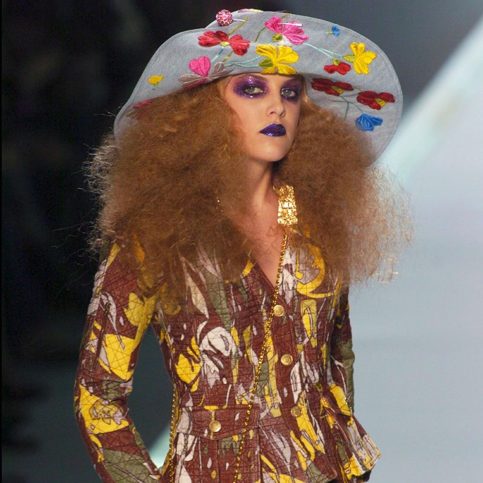 riley keough poses with her hand on a small purse while standing on a runway, she wears a hat with embroidered flowers, a floral patterned jacket, and khaki shorts