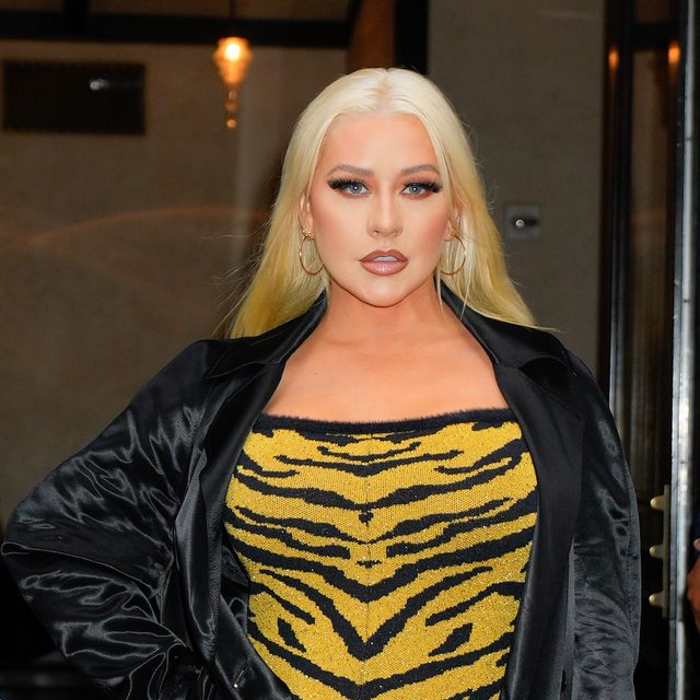 christina aguilera looks at the camera with one hand on her hip, she wears a black velvet jacket over a yellow and black tiger stripped dress