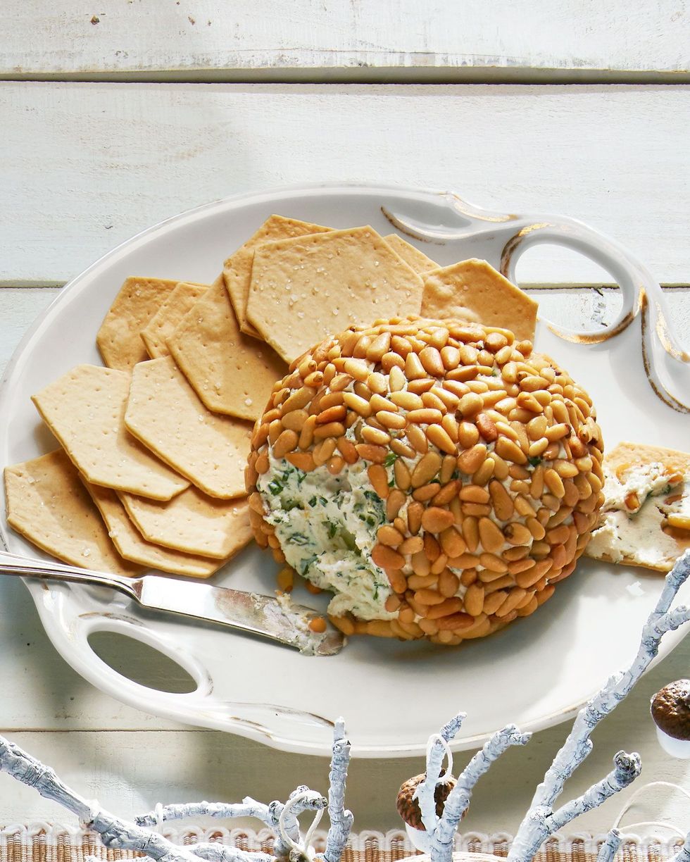 herbed cheese ball covered in toasted pine nuts on a white serving tray with handles and crackers for serving
